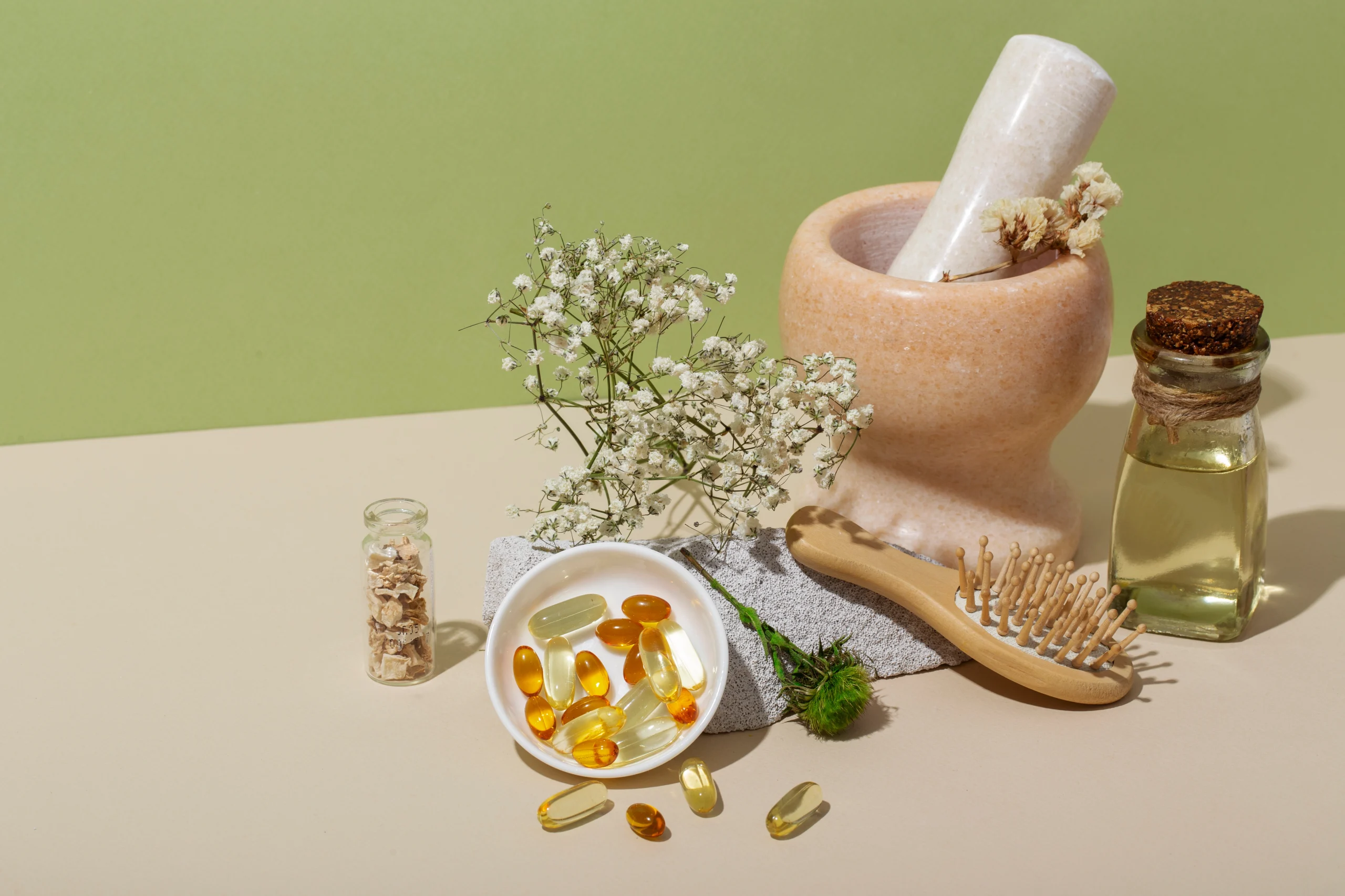 Flat lay of natural health items on a beige surface: mortar and pestle, oil bottle, dried flowers, and a dish of yellow-orange capsules with a wooden comb nearby